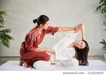 Masseuse making thai yoga massage. Woman therapist doing traditional massage treatment, stretching female patient's body. Client sitting on mat, enjoying hand massage in calm atmosphere. Masseuse making thai yoga massage. Woman therapist doing traditional massage treatment, stretching female patient's body. Client sitting on mat, enjoying hand massage in calm atmosphere. 123028859