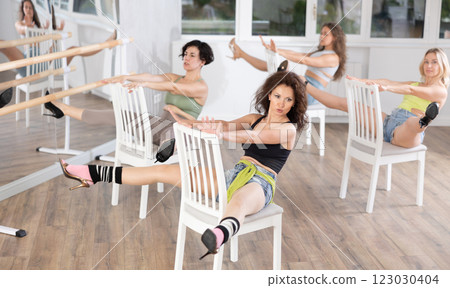 Beautiful young woman practicing strip plastic movements while sitting on chair and graceful leg stretch workout with group of athletic women at dance class Beautiful young woman practicing strip plastic movements while sitting on chair and graceful leg stretch workout with group of athletic women at dance class 123030404