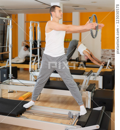 Man stands on reformer and holds ring, hoop in his hands, performs exercises to balance Man stands on reformer and holds ring, hoop in his hands, performs exercises to balance 123030578