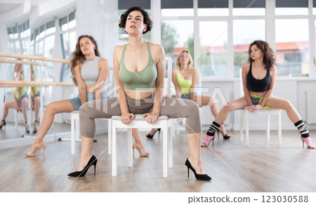 Pretty female choreographer demonstrating sensual and modern high heels choreography sitting on chair for sporty women during group class of strip plastic in studio 123030588
