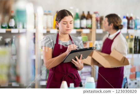 Female manager makes notes on an electronic tablet - conducts an inventory. In background, male salesperson places goods on shelves Female manager makes notes on an electronic tablet - conducts an inventory. In background, male salesperson places goods on shelves 123030820