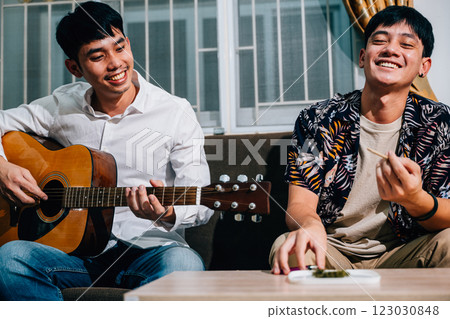 Two Asian men are sitting on a couch, one playing a guitar and the other smoking a cigarette while sitting on sofa in relaxing time at home. smoking marijuana or cannabis Two Asian men are sitting on a couch, one playing a guitar and the other smoking a cigarette while sitting on sofa in relaxing time at home. smoking marijuana or cannabis 123030848