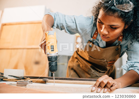 Carpenter woman using a power drill to secure a piece of wood in a workshop, focusing on precision in furniture making. Surrounded by tools and wood pieces, highlighting skill and craftsmanship Carpenter woman using a power drill to secure a piece of wood in a workshop, focusing on precision in furniture making. Surrounded by tools and wood pieces, highlighting skill and craftsmanship 123030860