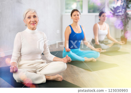 Elderly young women sitting on mats in Lotus Position Padmasana during yoga stretch joint workout in fitness center Elderly young women sitting on mats in Lotus Position Padmasana during yoga stretch joint workout in fitness center 123030993