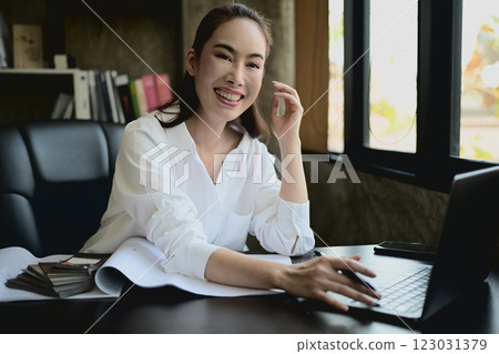 A confident Asian female architect boss sits at her desk, smiling warmly at the camera. 123031379