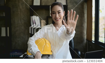A confident female engineer boss stands in her office, smiling and showing her palm at the camera. 123031404