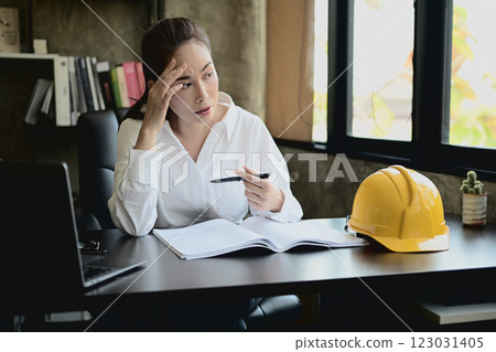 A stressed, tired Asian female architect or engineer boss sits at her desk in her private office. 123031405