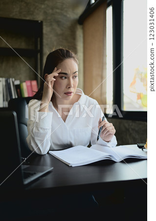 A stressed Asian female architect or engineer boss sits at her desk in her private office. 123031406