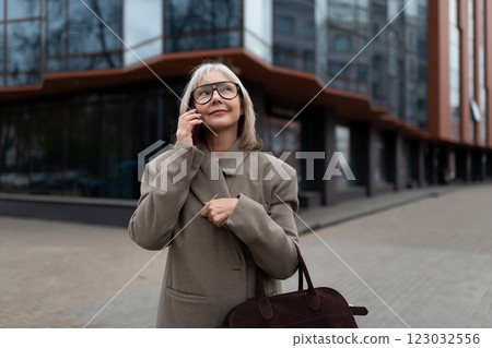Senior businesswoman talking on her phone while standing outside a modern office building in the Senior businesswoman talking on her phone while standing outside a modern office building in the 123032556