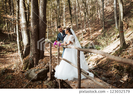 Beautiful bride and an elegant groom standing on forest bridge in nature, surrounded by a trees. 123032579