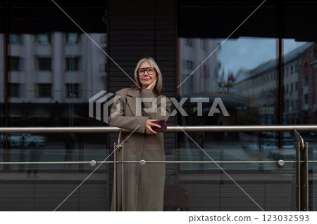 An older woman stands thoughtfully by a railing outside a modern building on a cloudy day in the An older woman stands thoughtfully by a railing outside a modern building on a cloudy day in the 123032593