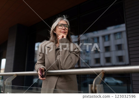 Elderly businesswoman pauses thoughtfully outside her office building during a sunny day Elderly businesswoman pauses thoughtfully outside her office building during a sunny day 123032596