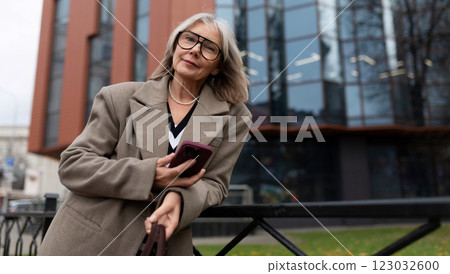 Senior businesswoman standing outside a modern office building, confidently using her smartphone 123032600