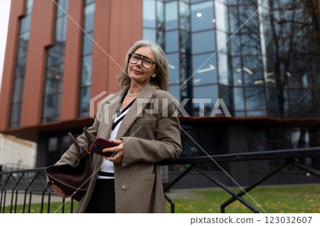 Confident elderly businesswoman holding a purse while standing outside a modern office building Confident elderly businesswoman holding a purse while standing outside a modern office building 123032607