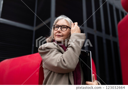An older woman standing confidently outdoors while adjusting her glasses with a laptop tucked under 123032664