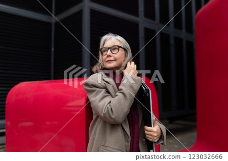 Stylish elderly woman with glasses and a folder poses confidently against vibrant red architectural Stylish elderly woman with glasses and a folder poses confidently against vibrant red architectural 123032666