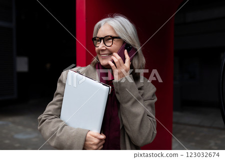 Middle-aged businesswoman smiling while talking on her phone, holding a folder outside an office 123032674