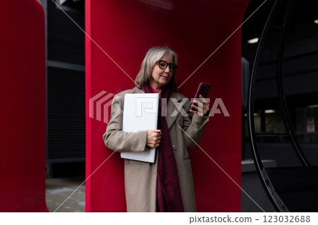 An older woman checks her phone while holding a laptop in a modern urban setting during the daytime An older woman checks her phone while holding a laptop in a modern urban setting during the daytime 123032688