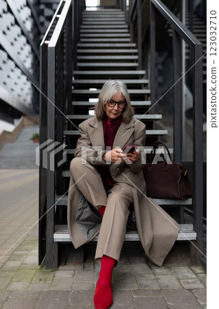 Middle-aged businesswoman in a tailored beige suit sits on stairs checking her phone in an urban 123032710
