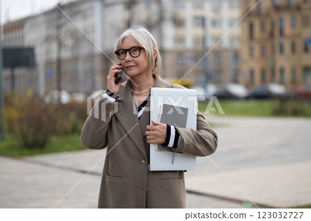 Senior businesswoman speaking on the phone while holding a notebook and documents on a city street Senior businesswoman speaking on the phone while holding a notebook and documents on a city street 123032727