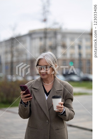 Senior businesswoman using her smartphone while holding a coffee cup on a city street in a Senior businesswoman using her smartphone while holding a coffee cup on a city street in a 123033406