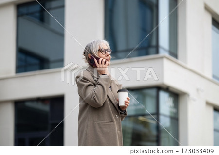 Older woman talking on the phone while holding a coffee cup outside a modern building on a sunny day 123033409