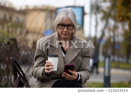 Older woman sitting on a bench outdoors, sipping coffee and browsing on her smartphone during a cool Older woman sitting on a bench outdoors, sipping coffee and browsing on her smartphone during a cool 123033414