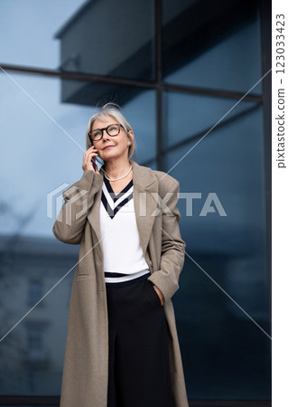 Senior businesswoman in a stylish coat makes a phone call outside a modern office building during 123033423