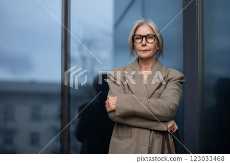 An older woman in stylish glasses stands confidently outside a modern building on a cloudy day 123033468
