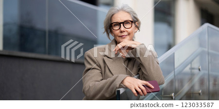 An older woman dressed in a beige coat stands thoughtfully by a modern building while holding a An older woman dressed in a beige coat stands thoughtfully by a modern building while holding a 123033587