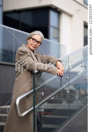Middle-aged businesswoman in glasses leans on glass railing at a modern urban building on a sunny Middle-aged businesswoman in glasses leans on glass railing at a modern urban building on a sunny 123033597