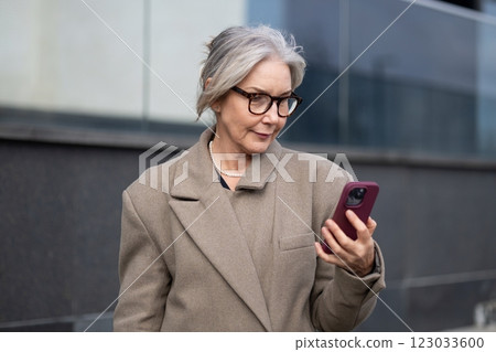 Elderly businesswoman checks her smartphone while standing outside a modern office building on a Elderly businesswoman checks her smartphone while standing outside a modern office building on a 123033600