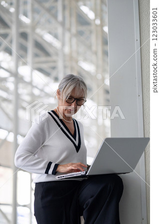 Elderly businesswoman working on her laptop outside a modern office building during the day Elderly businesswoman working on her laptop outside a modern office building during the day 123033601
