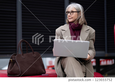 Senior businesswoman working outside on a laptop, dressed in a neutral suit, enjoying a break during 123033752