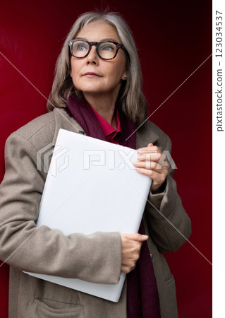 Stylish elderly woman with glasses confidently holding a folder against a striking red backdrop in Stylish elderly woman with glasses confidently holding a folder against a striking red backdrop in 123034537