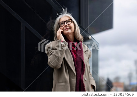 Elderly businesswoman talking on the phone outside her office building during a cloudy day Elderly businesswoman talking on the phone outside her office building during a cloudy day 123034569