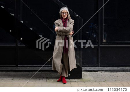 Stylish elderly woman in a long coat and red boots stands confidently against a modern urban Stylish elderly woman in a long coat and red boots stands confidently against a modern urban 123034570