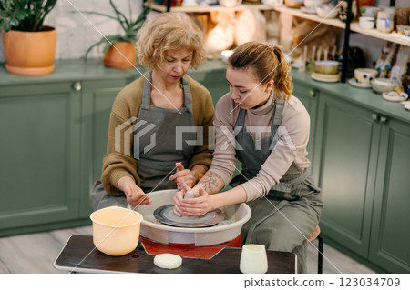 Mentor guiding elderly student in sculpting pottery on electric spinning wheel Two generations of women participate in hands-on ceramics training, shaping clay on a wheel 123034709