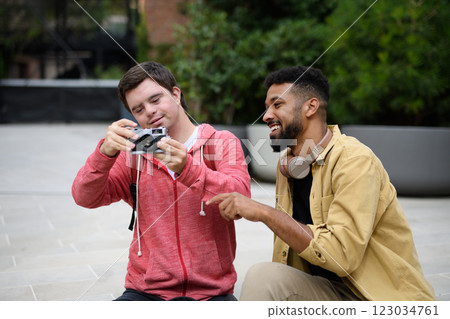 Young man with Down syndrome and mentoring friend spending time together, using camera outdoor. 123034761