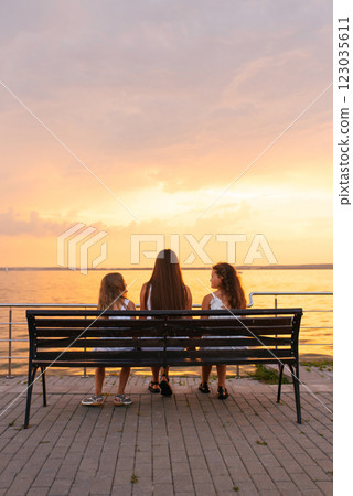 Mother sits on bench with two young girls near body of water, and the sky is a beautiful orange color. Concept of peace and relaxation 123035611