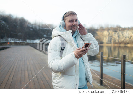 Smiling young man is walking near a lake on the forest, wearing headphones and using a mobile phone. 123036424