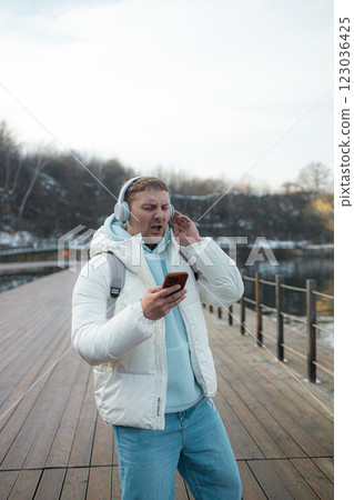 Smiling young man is walking near a lake on the forest, wearing headphones and using a mobile phone. 123036425