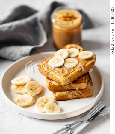 Peanut butter toast with banana on a plate on a light background with jar and napkin close up. Peanut butter toast with banana on a plate on a light background with jar and napkin close up. 123036833