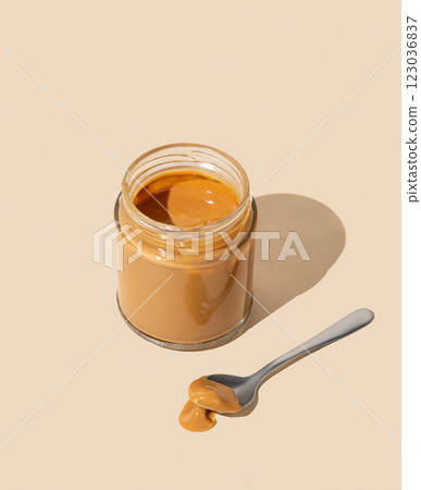 Peanut butter or caramel sauce in glass jar on isolated beige background with spoon and shadow. 123036837