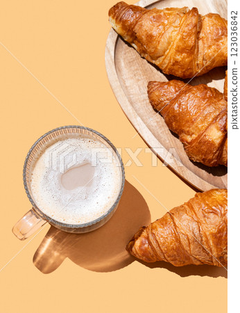 Cappuccino or latte coffee in a cup on an orange background with fresh croissants on a wooden plate. 123036842