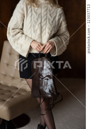 A girl in a warm sweater, a black short skirt and nylon tights is posing with a brown backpack. 123037713