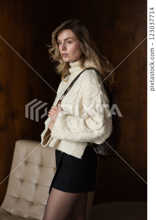 Woman in a warm sweater, a black short skirt and nylon tights is posing with a brown backpack. 123037714