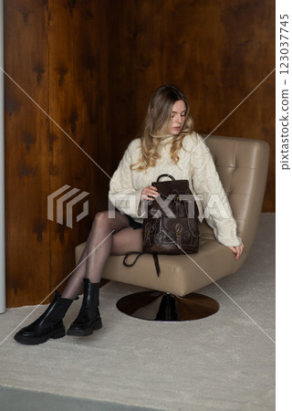 Woman in a warm sweater, a black short skirt and nylon tights is posing with a brown backpack. 123037745