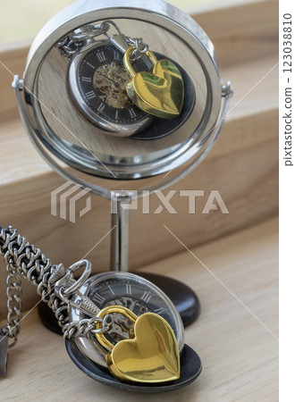 Golden heart shaped love padlock lap over A retro pocket watch with reflection in a circular mirror on wooden desk. Golden heart shaped love padlock lap over A retro pocket watch with reflection in a circular mirror on wooden desk. 123038810