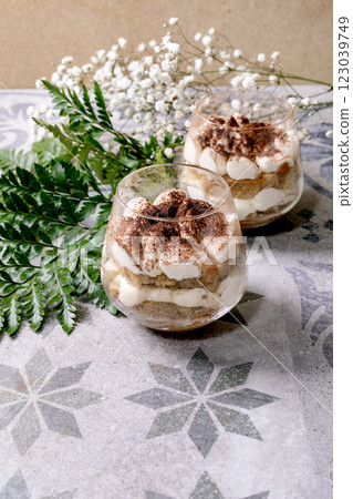 Classic Italian tiramisu in two glasses. Layers of sponge cake and mascarpone cream with cocoa powder. Placed on gray-blue patterned tile. White babys breath flowers, green leaves in background. 123039749